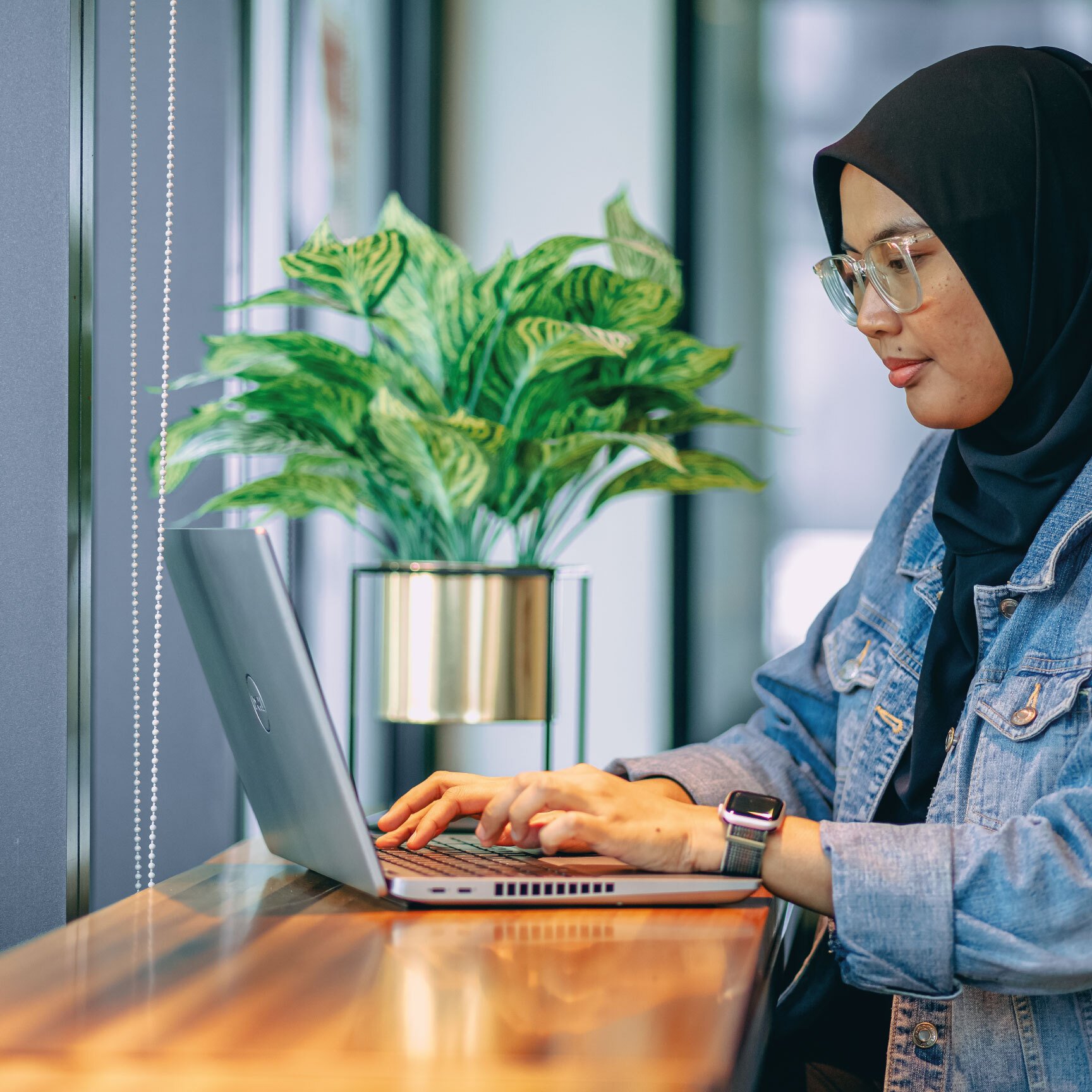 woman with a head scarf and jean jacket sits at table working on laptop