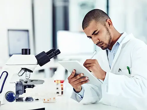 man in white medical coat taking notes on his tablet near a microscope clinic trials