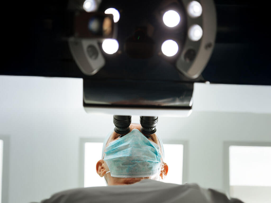 Medical professional using microscope in clinical setting under bright examination lights.