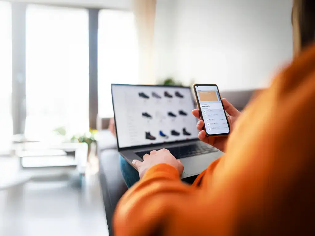 Person browsing online retail platform on laptop while reviewing data on smartphone.