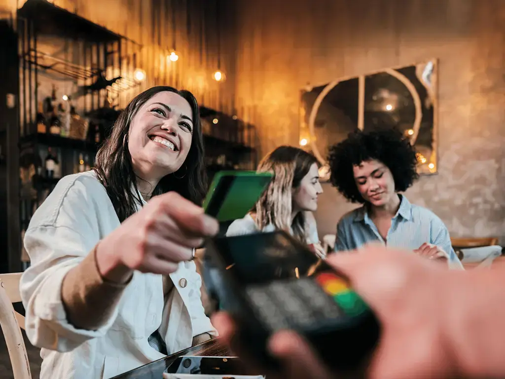 Smiling customer makes a contactless payment with a card at a restaurant while sitting with friends.