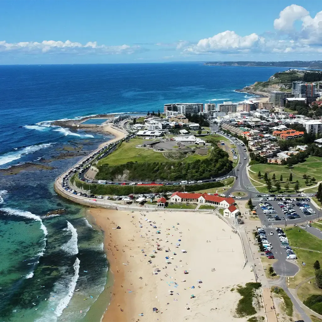 Aerial view of Newcastle, Australia, showcasing a sandy beach, ocean waves, green parks, and urban cityscape.