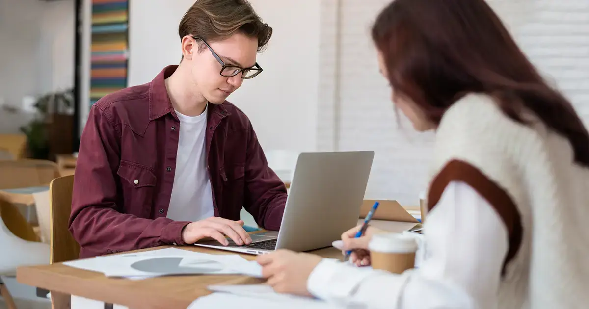 two people sit at a desk with a laptop, coffee and some paperwork