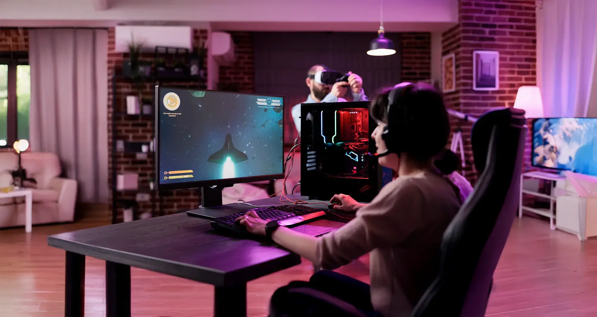 Person playing a space-themed video game on a desktop PC while another uses a VR headset in a gaming room setup.