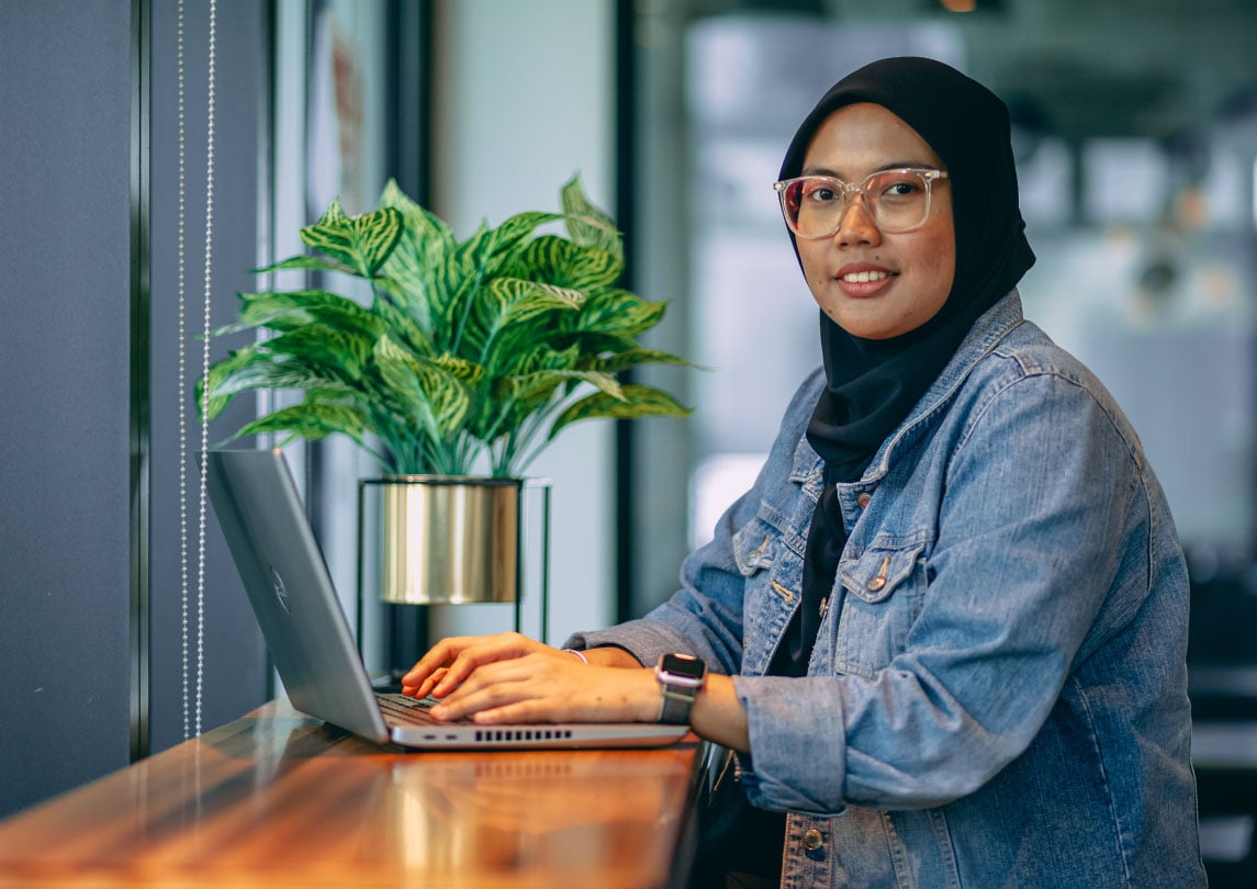 woman in a head scarf and jean jacket sits typing on her laptop at a counter