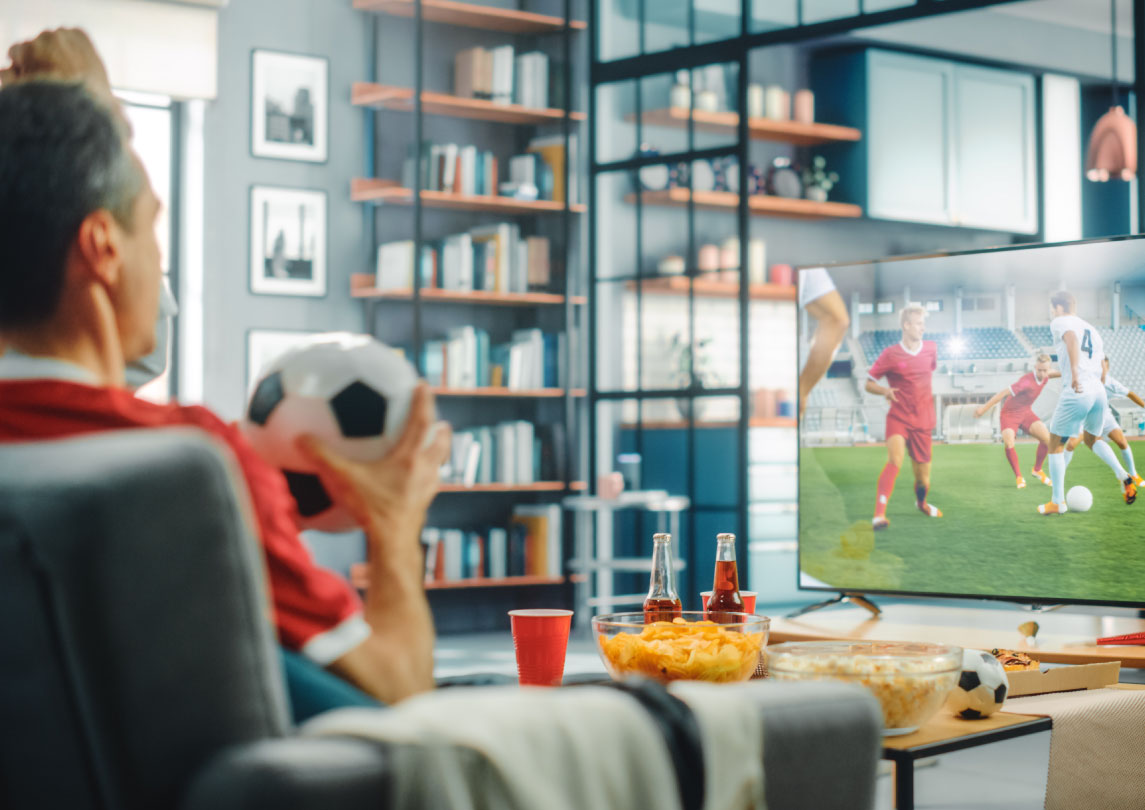 A man watching football game in front of his TV in his living room with a football held in his hands