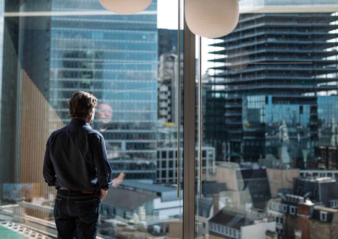 man in a navy collared shirt and jeans stands looking out a window at city skyscrapers